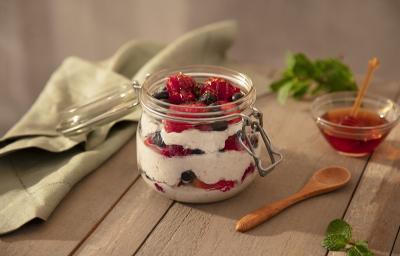 Foto da receita de overnight oats de frutas vermelhas servida em um pote de vidro hermético sobre uma mesa de madeira. Ao lado há um pano verde, uma colher de madeira e um pote de vidro com mel