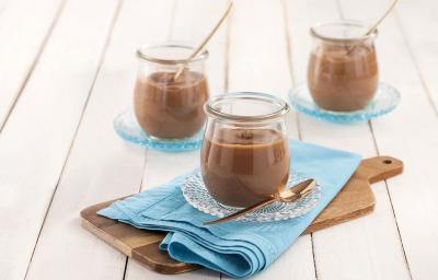 Fotografia em tons de azul em uma bancada de madeira branca, um pano azul turquesa, uma tábua de madeira e um potinho de vidro com a mousse de chocolate dentro dele. Ao fundo, mais potinhos com mousse.