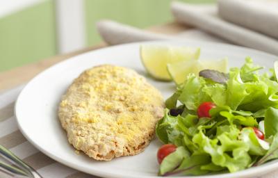 Fotografia em tons de verde em uma bancada de madeira com um pano branco com listras beges e um prato oval branco com o filé de frango empanado e salada verde dentro dele.