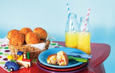 Fotografia em tons de azul e vermelho em uma mesa antiga vermelha com pratos coloridos, cesta de vime com os lanchinhos de frango sem leite e sem ovo e garrafas de vidro sobre a mesa.