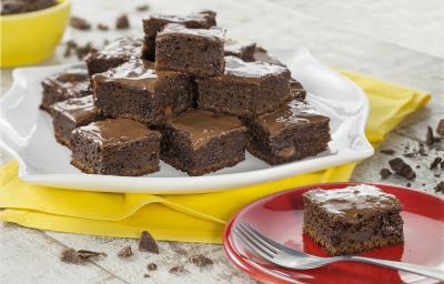 Fotografia em tons de chocolate, branco e amarelo, com prato branco com pedaços de bolo de chocolate sobre guardanapo amarelo, prato vermelho com porção de bolo e um garfo, no alto pote amarelo com chocolate, sobre bancada clara com pedaços de chocolate.