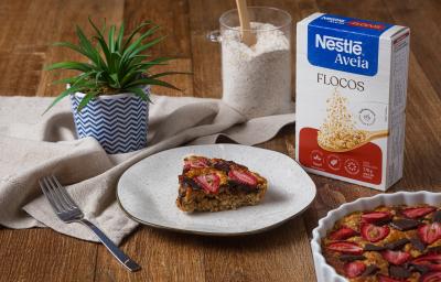 Fotografia de um prato branco com borda preta, ao lado de uma embalagem de aveia flocos, sobre uma mesa de madeira. Sobre o prato tem uma fatia do bolo de aveia com morango e chocolate.