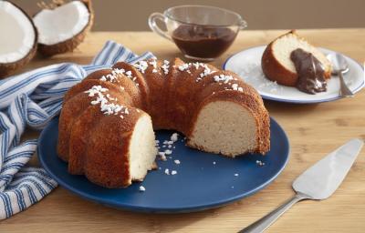 Foto da receita de bolo de leite de coco cremoso caseiro servida em uma porção com uma fatia cortada sobre um prato de porcelana azul escuro com a fatia sobre um prato de sobremesa branco com uma cobertura de chocolate e ao lado um paninho listrado