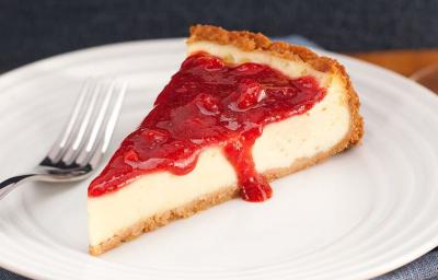 Fotografia em tons de branco e vermelho, no centro na foto está uma fatia da torta coberta com geleia de morango, ao lado está um garfo e embaixo do prato um guardanapo azul.