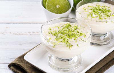 Foto aproximada da receita de Mousse de Limão em tom claro, em um pote redondo de vidro e decorado com raspas de limão. Tudo sobre um prato branco e um tecido marrom escuro. No rodapé há uma faixa verde com dizeres.