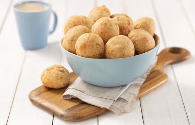 Foto em tons de bege com os pães de queijo em um bowl azul claro sobre um paninho cinza em cima de uma tábua de madeira, com uma unidade à frente do bowl para decorar. No fundo como decoração há uma xícara azul de café.