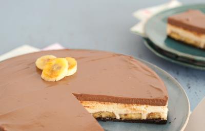 Fotografia em tons azul em uma mesa de madeira azul, com um pano branco com rosa florido, um prato azul raso redondo com a torta mousse de doce de leite e banana.