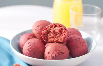 Fotografia em tons de azul e rosa em uma mesa branca com um pano azul ao lado e um prato branco fundo ao centro com os pães de beterraba. Ao fundo, uma jarra com suco de laranja e um copo de vidro.