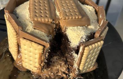 Fotografia de um bolo de chocolate com brigadeiro e farinha láctea polvilhada. Ao redor do bolo tem chocotrio de amendoim grudado. O bolo está sobre um prato de vidro preto raso.