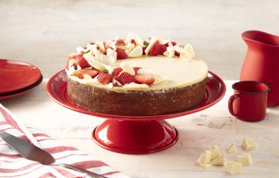 Fotografia em tons de vermelho em uma bancada bege com um paninho listrado branco e vermelho e ao centro um suporte vermelho com o bolo de três leites com ganache de chocolate branco. Ao lado, pedaços de chocolate branco, uma caneca e uma jarra vermelhas.