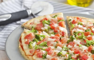 fotografia em tons de branco de uma bancada vista de frente, contém um prato redondo e branco com uma pizza com pedaços de tomates e brócolis ao fundo uma jarra com azeite.