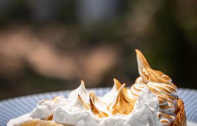 Fotografia de uma fatia de torta de cupuaçu feita com creme de leite, e por cima tem um merengue maçaricado. A fatia está em um prato de vidro branco com bordas cor na cor azul. Ao fundo da foto tem algumas árvores.