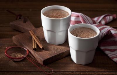 Foto dois copos brancos com chocolate quente dentro em cima de uma tábua de madeira com canela em pau e em pó decorando a mesa