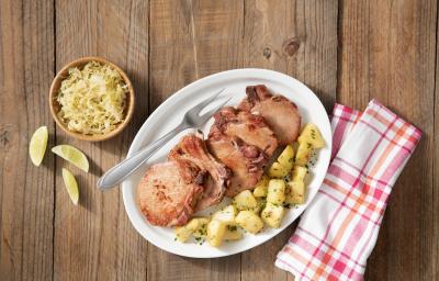 Fotografia em tons de branco de marrom, com bancada de madeira vista de cima, ao centro travessa branca contendo bisteca suína, batata picada e um garfo, no entorno potinho com repolho e fatias de limão e guardanapo quadriculado em branco e vermelho.