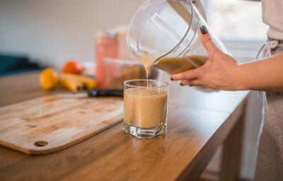 Fotografia de uma moça colocando uma bebida de banana e maçã feita no liquidificador em um copo de vidro pequeno, que está sobre uma mesa de madeira. Ao fundo, uma tábua de madeira e frutas, como bananas mamão e maçã.