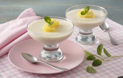 Fotografia em tons de rosa e amarelo de uma bancada, ao centro um paninho quadriculado rosa e branco, um prato rosa com uma taça com o mousse e pedacinhos de abacaxi e folha de hortelã. Ao lado uma taça igual e folhas de hortelã.