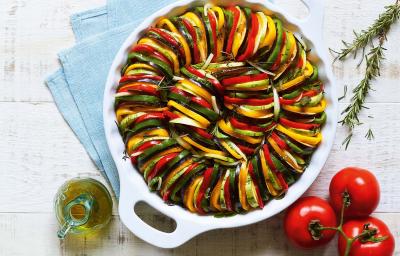 Fotografia em tons de verde, amarelo e vermelho de uma bancada branca com três tomates, uma jarrinha de azeite, ramos de alecrim e um recipiente branco redondo com alças com camadas intercaladas de abobrinha, berinjela e pimentões.