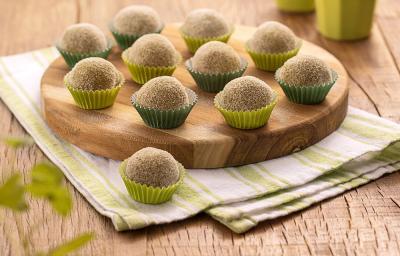 Foto da receita de Brigadeiro de Batata Doce. Na foto, observa-se brigadeiros em forminas nos tons de verde, sobre uma tábua redonda de madeira e um guardanapo listrado.