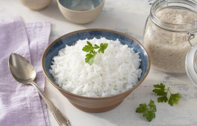 Fotografia em tons de lilás em uma bancada de madeira clara, um pano lilás e um pote marrom redondo fundo com o arroz basmati dentro dele. Ao lado, um pote hermético com arroz cru e uma colher de servir.