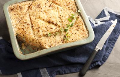 Fotografia em tons de dourado e azul, assadeira com a torta de pão de forma gratinada com queijo e folhas verdes, sobre guardanapo azul com listras brancas, faca ao lado, tudo sobre toalha cinza.