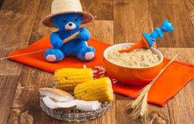 Fotografia em tons de laranja e azul de uma bancada de madeira com um paninho laranja e um potinho com uma papinha dentro. A frente uma espiga de milho ao meio, pedaços de coco e atrás um ursinho de pelúcia azul com um chapéu de palha.