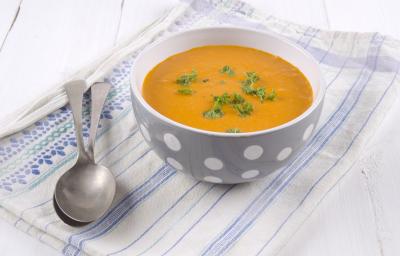 fotografia em tons de branco, laranja e azul de uma bancada branca vista de cima. Contém um pano branco com listras azuis, por cima um recipiente redondo cinza com bolinhas brancas e dentro a sopa. Ao lado do recipiente duas colheres.