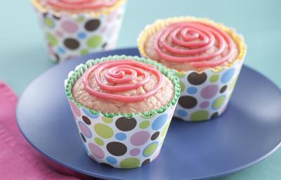 Fotografia em tons de azul e rosa em uma mesa de madeira azul, um pano rosa, um prato azul redondo com dois cupcakes de morango com aveia em forminhas coloridas.