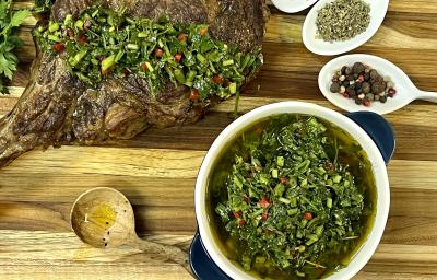 Foto da receita de Chimichurri. Observa-se um carré de cordeiro com o molho por cima sobre uma tábua de madeira.