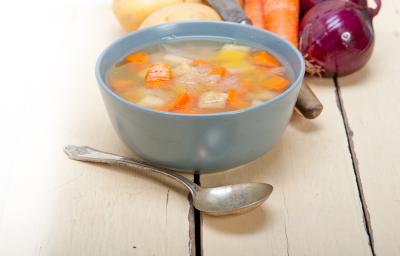 Fotografia de uma sopa de batata, cenoura e chuchu, dentro de um recipiente de vidro na cor azul. Na frente, uma colher de sopa, e ao fundo, cebolas roxa, batatas e cenoura, sobre uma mesa de madeira.