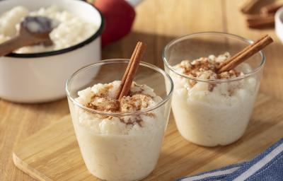 Fotografia em tons de branco em uma bancada de madeira marrom. No centro, dois copos contendo arroz doce. Ao fundo, uma panela branca contendo arroz doce e algumas canelas em pau espalhadas.