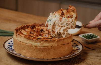 Fotografia em tons de marrom de uma bancada de madeira com um prato raso branco com detalhes azul sobre ele um quiche de frango com um pedaço sendo cortado. Ao fundo um potinho branco quadrado com cebolinha e uma cebola.