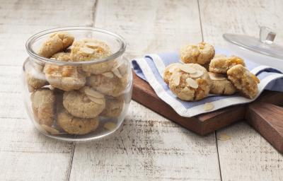 Fotografia em tons de azul em uma bancada de madeira clara com um pano azul sobre uma tábua de madeira com alguns cookies de amêndoas e damasco apoiados nele. Ao lado, um pote grande de vidro com mais cookies dentro.