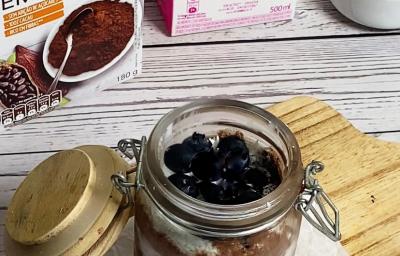 Foto da receita de pudim de chia servida em um pote hermético de vidro sobre uma tábua de madeira em cima de uma mesa de madeira branca. Ao fundo há embalagens de nature's heart cacau em pó e bebida vegetal de amêndoa