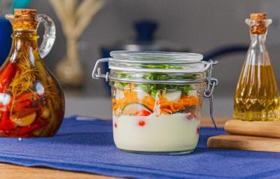 Fotografia em tons de azul com um pote de vidro ao centro. Dentro do pote existe uma receita de salada rúcula com cenoura, pepino, tomate cereja com molho caesar