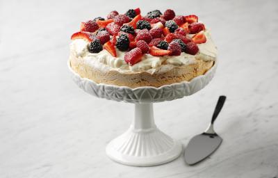 Fotografia em tom claro, com uma torta recheada com suspiro e frutas por cima, polvilhadas com açúcar de confeiteiro. A torta está apoiada sobre um prato alto branco decorativo, ao lado, uma espátula prata para bolo, tudo sobre uma mesa de mármore branca.