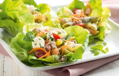 Fotografia em tons de branco e verde, ao centro salada de folhas verdes com macarrão, legumes e mozarela, sobre guardanapo rosa e bancada de madeira em tons de branco.