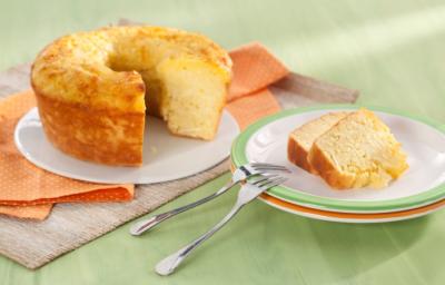 Fotografia em tons de laranja e verde em uma mesa de madeira verde com um pano laranja de bolinhas branca. Ao centro, um prato raso branco com o bolo de macaxeira cortado. Ao lado, um prato branco com borda verde e duas fatias do bolo.