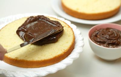 Foto de um bolo aberto ao meio em cima de um prato branco sendo preenchido com creme de chocolate por cima