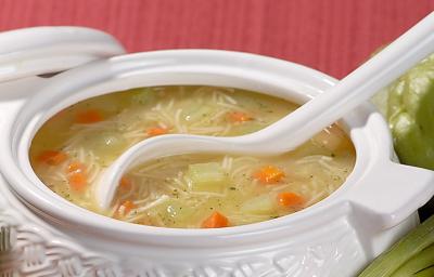 Fotografia em tons de branco, vermelho e verde, com sopeira branca ao centro com sopa de legumes, macarrão e um concha branca, tendo em volta chuchu, cebolinha, cenoura, cebola, salsa e uma tampa branca, tudo sobre bancada vermelha.