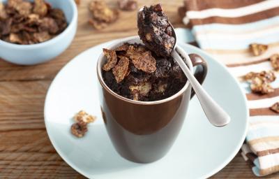 Fotografia em tons de marrom em uma bancada de madeira com um pano listrado em marrom, azul claro e bege ao lado e ao centro, um prato branco com uma xícara marrom em cima dele e o bolo de caneca de chocolate dentro, decorado com os cereais Nesfit.