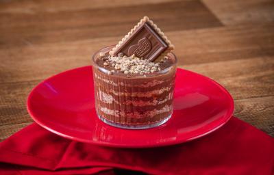 Fotografia de um copo de vidro pequeno com camadas de chocolate alpino derretido e chocobiscuit triturado. Por cima tem choco biscuit triturado e um biscoito inteiro apoiado para decorar. O copo está sobre um prato e uma toalha de mesa vermelha.