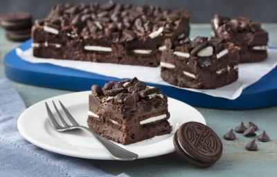 Fotografia em tons de azul em uma bancada de madeira cinza, um pano azul, uma tábua de madeira azul com um papel manteiga e o brownie com recheio de Biscoito Negresco dentro e em cima do papel. Ao centro, um prato branco redondo com um pedaço do brownie.