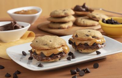 Fotografia em tons de amarelo em uma bancada de madeira com um recipiente branco ao centro com dois cookies recheados com doce de leite e creme de avelã. Ao fundo, potinhos com gotas de chocolate, doce de leite e uma tábua com mais cookies.