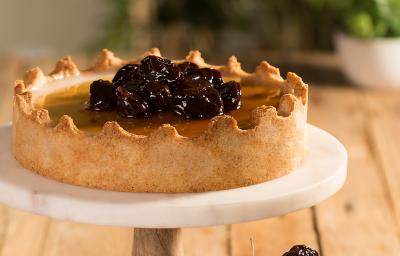 Fotografia em tons de marrom em uma bancada de madeira com um suporte de bolo branco com a torta manjar em cima. Ao lado, um potinho de vidro com ameixas secas pretas.