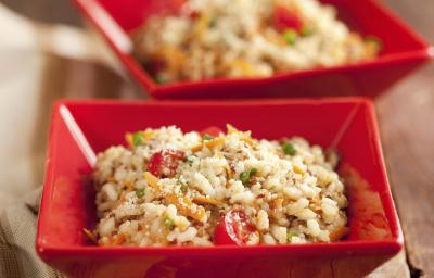 Fotografia em tons de vermelho, marrom e branco, com dois potinhos quadrados contendo um arroz colorido  e queijo ralado, ao lado guardanapo em tons de branco e marrom, tudo sobre bancada de madeira rústica.