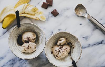 Fotografia em tons de amarelo, branco, azul e marrom de uma mesa branca e azul vista de cima, 2 recipientes redondos contém bolas de sorvete com pedaços de chocolates e cada um tem uma colher para servir. Ao fundo uma banana picada e pedaços de chocolate