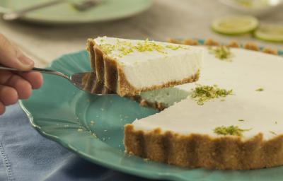 Fotografia em tons de verde, azul e branco de uma bancada branca com um paninho azul, sobre ele um prato verde redondo com uma torta de limão e uma espátula retirando uma fatia de torta. Ao fundo fatias de limão e um prato verde redondo.