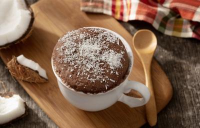 fotografia em tons de marrom e branco de uma bancada marrom vista de cima, contém uma tábua de madeira com uma xicara branca com bolo de prestígio e coco ralado por cima para decorar