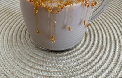 Fotografia em tons de marrom em uma bancada com um pano bege e uma xícara de vidro com a bebida de cappuccino de caramelo salgado dentro. Xícara decorada com calda de caramelo e biscoitos Tostines Especiarias