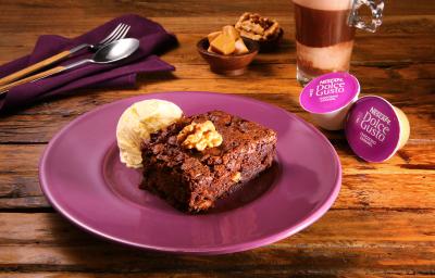 Fotografia em tons de roxo em uma bancada de madeira escura, um prato redondo roxo com o brownie com uma bola de sorvete de creme ao lado. Ao fundo, um pano roxo com uma colher e um garfo e ao lado uma xícara com o chococino caramel Dolce Gusto.